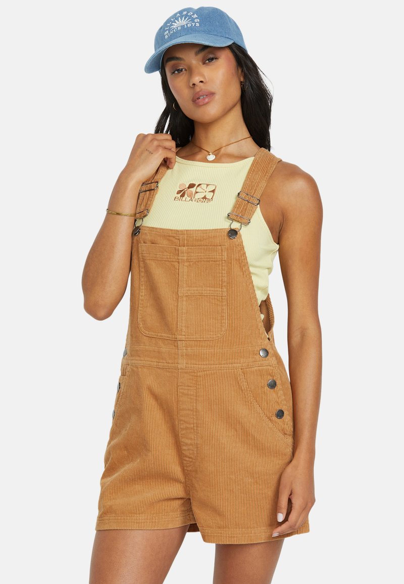 Salopette di corduroy di un caldo colore tan, con una parte anteriore a bavaglino, dettagli con bottoni laterali e pantaloni corti rilassati. Abbinata a una maglietta verde chiaro.