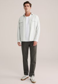 Jongeman in een wit gestreept overhemd over een wit T-shirt, donkere grijze broek en witte sneakers met gum-zolen, staand voor een beige achtergrond.