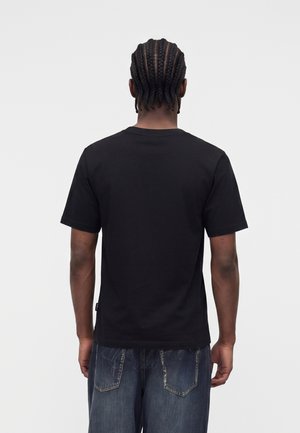 Mężczyzna z warkoczami, ubrany w zwykłą czarną koszulkę z krótkim rękawem i wyblakłe niebieskie dżinsy, stoi tyłem do kamery.