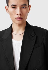 Blazer nero con rever a lancia, spalle strutturate e una tasca sul petto. Indossato sopra una camicia bianca strutturata. Design minimalista.