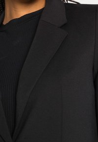 Blazer noir avec des revers structurés, coupe ajustée et texture lisse, superposé à une chemise noire côtelée, offrant un look élégant et monochrome.