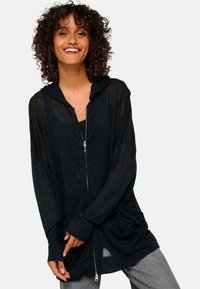 Sweat à zip noir en tissu léger avec une coupe décontractée, des manches longues et des poches avant, associé à un pantalon gris.