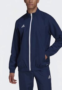 Laivastonsininen urheilutakki vetoketjulla edessä, elastisilla hihansuiden ja helman resoreilla. Ominaisuuksiin kuuluu valkoiset korostukset ja Adidas-logo vasemmassa rinnassa.