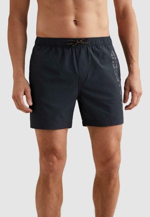 Man wearing black drawstring swim shorts with vertical logo on right thigh against plain background.
