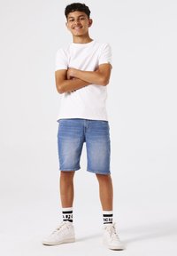 Camisa de algodón blanca, pantalones cortos de mezclilla azules, zapatillas blancas y calcetines con patrón en blanco y negro. El modelo posa con los brazos cruzados.