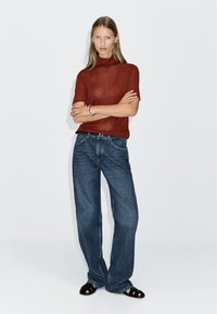 Rust-colored, short-sleeved knit top paired with loose-fit, dark blue denim jeans; worn with black, flat sandals.