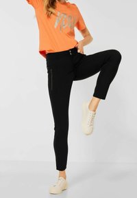Černé elastické kalhoty s úzkým střihem a bočními kapsami na zip, spárované s oranžovým tričkem se stříbrným grafickým textem, nošeny s bílými teniskami.