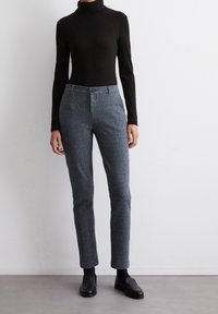 Un haut noir à col roulé associé à un pantalon gris pied-de-coq ajusté. Le pantalon présente une fermeture à bouton à l'avant et un design de jambe fuselée. Chaussures noires.