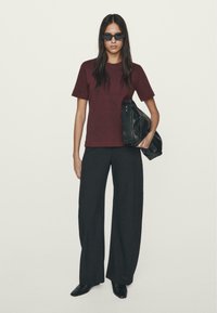 Maroon short-sleeve t-shirt with black wide-leg pants, holding a black bag. Model wears dark sunglasses and has long hair. Smooth fabric texture.