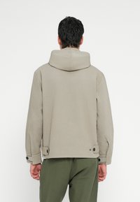 Bež jopa s kapuco z ohlapnim krojem, poudarjen z gumbi, zaprtimi manšetami in gladko teksturo, zasnovan za priložnostno nošenje.