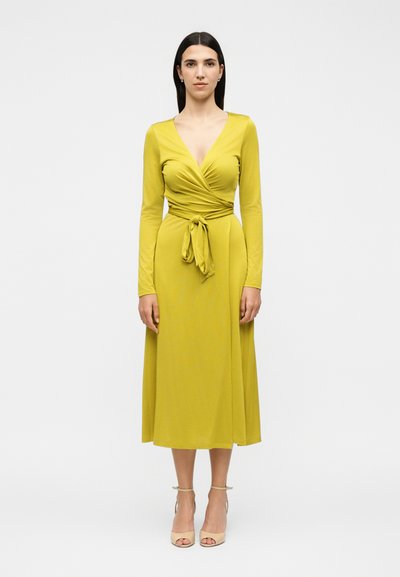 Woman standing in a long-sleeve, mustard yellow wrap dress tied at the waist, wearing beige open-toe heels against a plain white background.