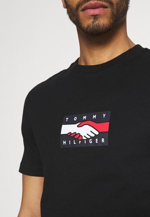 Zwart T-shirt met korte mouwen met een grafische print van twee handen die elkaar schudden in rood en wit tegen een marineblauwe achtergrond met de tekst "TOMMY HILFIGER".