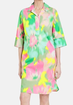 Femme portant une robe ample jusqu'aux genoux avec un motif floral abstrait coloré en vert, rose, jaune et bleu clair, avec des manches trois-quarts.