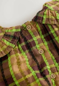 Braun-grün kariertes Hemd mit einem Rüschensaum und Holzknöpfen, das eine Mischung aus horizontalen und vertikalen Streifen aufweist. Weicher Stofftextur.