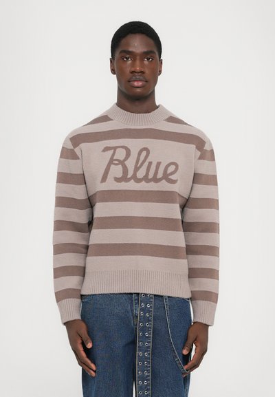 Bruine gestreepte trui met gebreide textuur, met het woord "Blauw" in lichtbruin. Ribbelkraag en -manchetten, gecombineerd met blauwe denim broek.