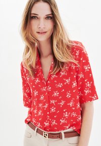 Röd kortärmad skjorta med mönster av blommor, knäppning framtill, lättviktig tyg, kombinerad med ett beige bälte och beige byxor.