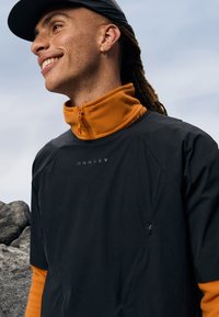 Oseba z nasmehom nosi črno kapo, zlate obroče, oranžno flis zapenjanje ter črno Oakley jakno, stoji blizu sivih kamnov pod oblačnim nebom.