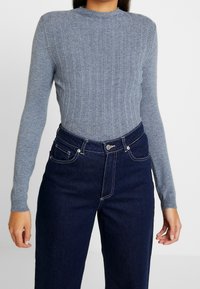 Maglione grigio a coste con scollo rotondo e maniche lunghe, abbinato a jeans in denim scuro a vita alta con cuciture bianche.