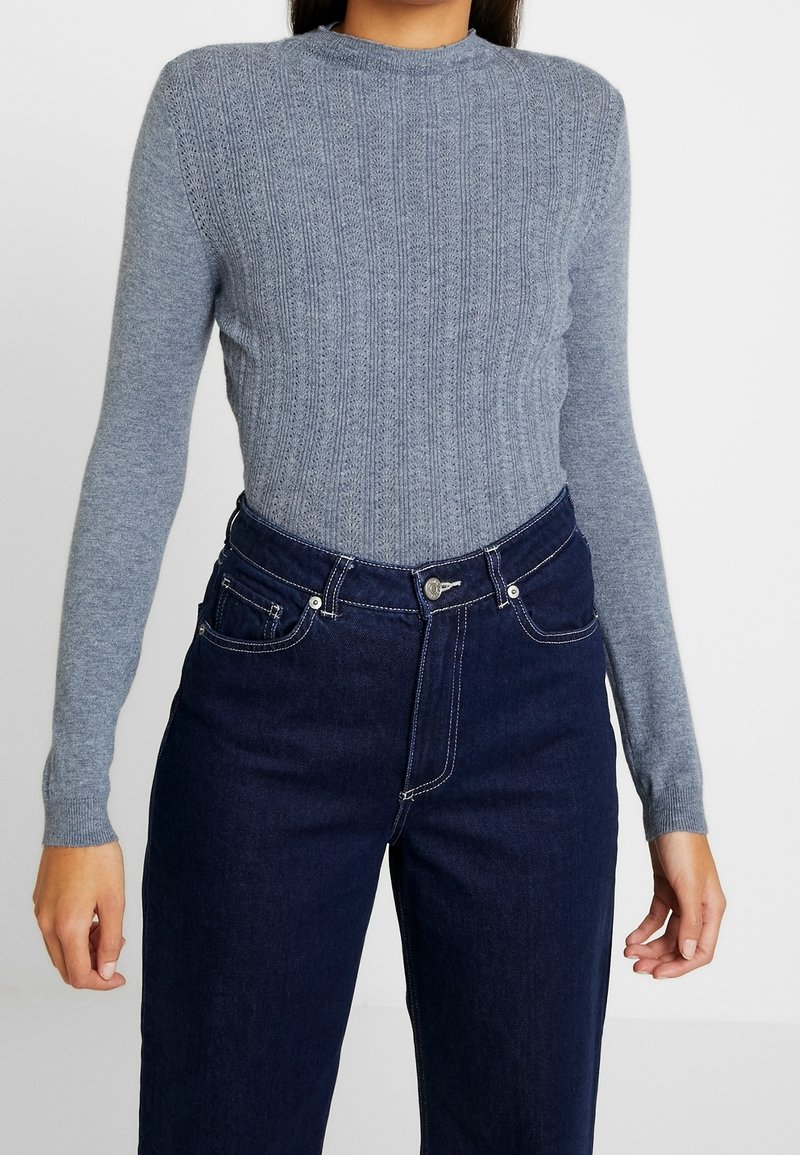 Maglione grigio a coste con scollo rotondo e maniche lunghe, abbinato a jeans in denim scuro a vita alta con cuciture bianche.