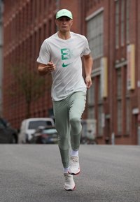 Hvit Nike T-skjorte med grønt logo, lysegrønne tights og hvite joggesko med røde detaljer. Løper på en bygate i bakgrunnen.