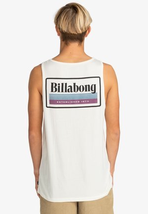 Osoba o krótkich blond włosach, ubrana w białą koszulkę bez rękawów z dużym logo Billabong na plecach oraz beżowe spodnie.