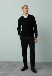 Schwarzer, gestrickter V-Ausschnitt-Pullover über einem weißen Kragenhemd, kombiniert mit schwarzen Hosen und schwarzen Schnürschuhen. Einfarbiger Hintergrund.