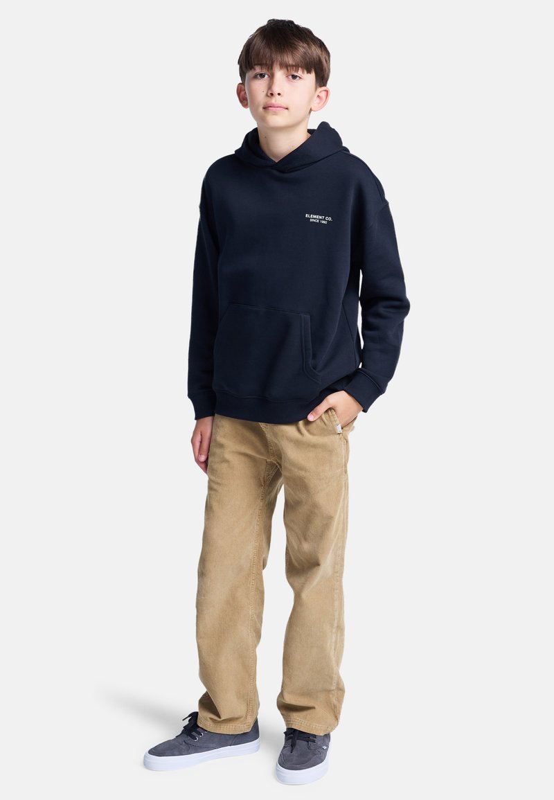 Felpa navy con tasca frontale e testo del logo bianco, abbinata a pantaloni in corduroy beige e sneakers grigie. Outfit semplice e casual.