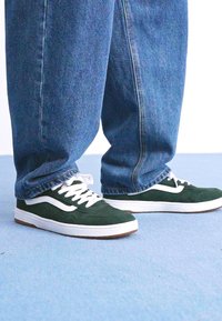 Pés a usar sapatilhas de camurça verde-escuro com atacadores e risca brancos, combinadas com uns jeans de ganga azuis largos num chão azul-claro.