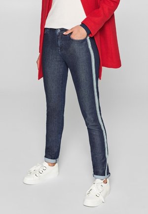 Person trägt dunkelblaue Jeans mit hellblauen Seitenstreifen, weiße Sneakers und einen roten Mantel mit Händen in den Taschen und steht vor einem einfarbigen Hintergrund.