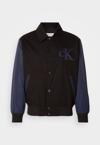 Black jacket with navy sleeves, button closure, and embroidered "cK" logo. Features a collared neck and elasticized cuffs for a fitted look.