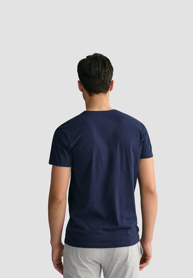 Uomo di spalle che indossa una maglietta blu navy a maniche corte tinta unita e pantaloni grigio chiaro su uno sfondo grigio chiaro uniforme.