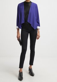 Lila öppen jacka över en svart texturerad topp, kombinerad med figurnära svarta byxor och blanka svarta ankelstövlar. Enkel svart kuvertväska i handen.