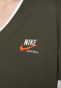 Personne portant un haut de sport en maille vert olive avec bordure blanche et logo Nike Sportswear blanc et orange sur la poitrine.