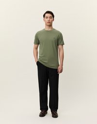 Mann som står iført olivengrønn kortermet T-skjorte, svarte bukser og brune sko mot en ensfarget lys bakgrunn.