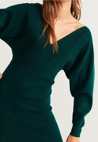 Robe en tricot d'un bleu sarcelle profond, avec un décolleté en V, des manches bouffantes longues et une taille cintrée. La texture est douce avec un motif côtelé.