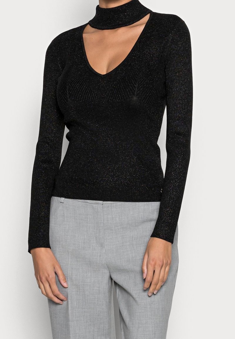 Femme portant un haut en maille noir à manches longues avec un col en V et un col ras du cou découpé, associé à un pantalon taille haute gris clair.