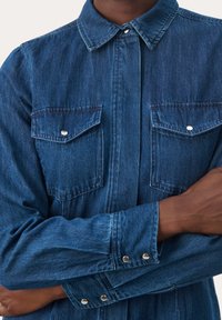 Tummansininen farkkupaita, jossa on suippo kaulus. Sitä koristavat kaksi rintataskua, hopeiset nepparit ja hihansuiden yksityiskohdat. Silkkinen pinta.