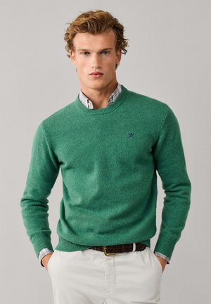 Hombre con jersey verde sobre camisa a cuadros, con pantalones blancos y cinturón marrón tejido, de pie con las manos en los bolsillos.