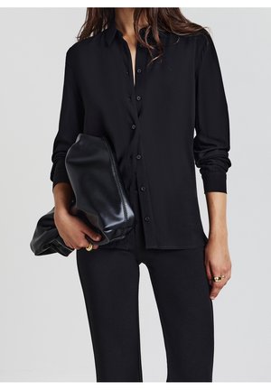 Femme portant une chemise noire boutonnée et un pantalon noir, tenant une grande pochette en cuir noir avec des bagues dorées aux doigts.