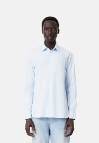 Camisa de manga larga azul claro con cuello abotonado, presenta rayas verticales blancas y un logo bordado en el pecho. Material de algodón.