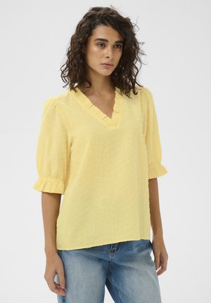 Woman with curly dark hair wearing a textured yellow V-neck blouse with ruffle sleeves and light blue jeans, standing against a plain background.