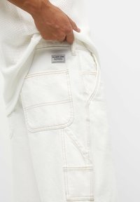 Pantalones cortos de carga blancos de algodón con costuras visibles, bolsillos laterales y una etiqueta de marca en la cinturilla trasera.
