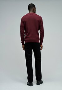 Homme debout de dos portant un pull texturé bordeaux, un pantalon noir et des chaussures noires, sur un fond gris clair uni.
