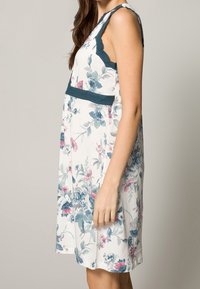 Robe à motifs floraux en blanc avec des fleurs bleues et roses. Présente des accents en dentelle le long de l'encolure et sous la poitrine. Design sans manches, longueur genoux.