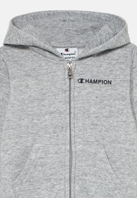 Pelēka izvelkamā kapuce no mīkstas auduma, ar kengūru kabatu un mazu melnu izšūtu "CHAMPION" logotipu uz krūtīm.