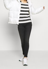 Bílá péřová bunda přes černobílé pruhované tričko s dlouhým rukávem, doplněná černými úzkými džínami a bílými teniskami s barevnými detaily.