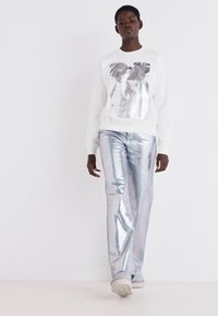 Felpa bianca con stampa grafica e testo, abbinata a pantaloni a zampa metallici argento lucido e sneakers di colore chiaro.