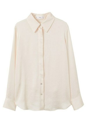 Femme portant une chemise blanche ample boutonnée, un jean large foncé, des sandales noires à talons, et tenant un petit sac à main noir avec une main dans la poche.