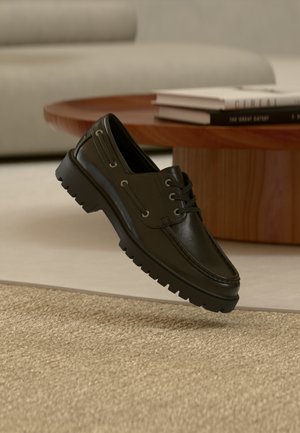 Zwarte leren veterboot die zweeft boven een beige, gestructureerd tapijt met een houten salontafel en een bank op de achtergrond in zachte focus.