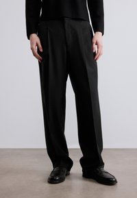 Μαύρο παντελόνι με ραφές, με λεία, γυαλιστερή επιφάνεια και πιέτες στο μπροστινό μέρος, συνδυασμένο με μαύρα μοκασίνια. Απλός σχεδιασμός, ίσια γραμμή παντελονιού.